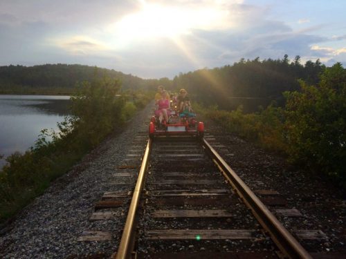 Two people ride a red rail bike on a scenic railway track beside a lake, with a sunset in the background.