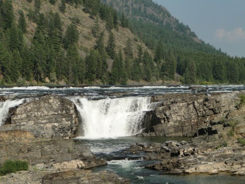 A scenic waterfall cascading over rocky cliffs, surrounded by lush green trees and mountains in the background.