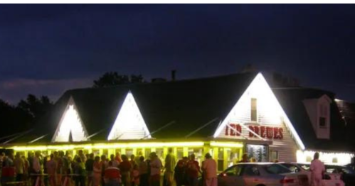 A crowded restaurant exterior at night, illuminated with bright lights and a long line of people waiting outside.