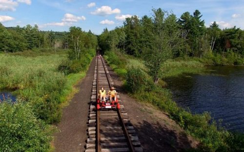 Two people ride a red rail cart along a train track surrounded by lush greenery and a calm body of water.