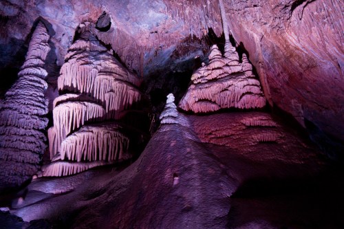 Colorful stalagmites and stalactites in a cave, illuminated by purple and pink lighting, creating a mystical atmosphere.