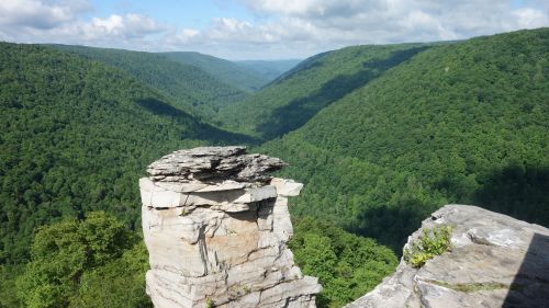A rocky outcrop overlooks a lush green valley surrounded by rolling hills under a partly cloudy sky.