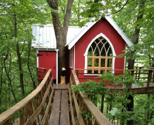 A charming red treehouse with a white roof, nestled among green trees, connected by a wooden walkway.