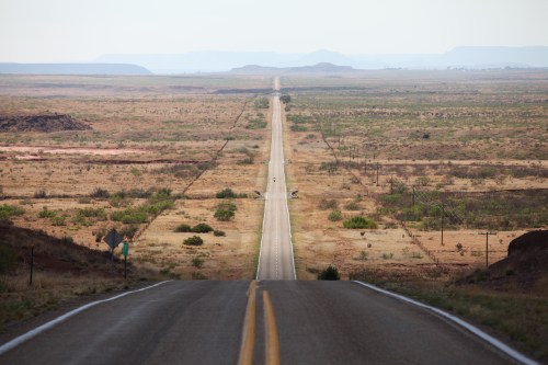 A long, straight road stretches through a vast, arid landscape under a clear sky. Mountains are faintly visible in the distance.