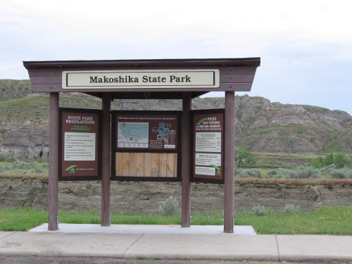 Signboard for Makoshika State Park with park regulations and a map, set against a backdrop of rugged hills.