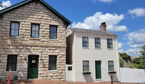 Two historic buildings side by side: a stone house with green trim and a white wooden house, under a blue sky with clouds.