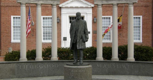 Statue of Thurgood Marshall in front of a brick building, flanked by flags and columns.
