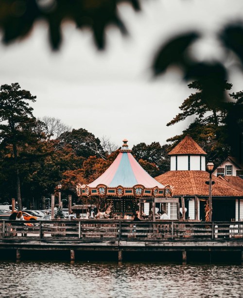 A colorful carousel near a lake, surrounded by trees and a quaint building, with people enjoying the ride.