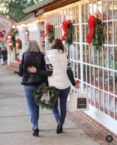 Two women walk together down a festive street, one holding a gift and the other a shopping bag, adorned with holiday decor.