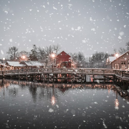A snowy scene of a quaint village by a lake, with a red barn and wooden bridge illuminated by soft lights.