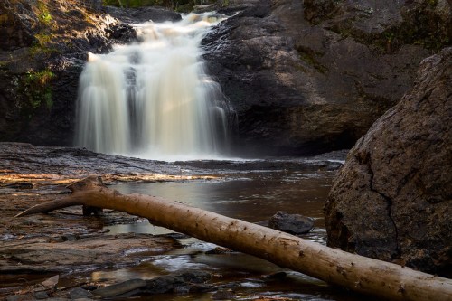 A serene waterfall cascades over rocks, with a fallen log resting on the riverbank in a tranquil natural setting.