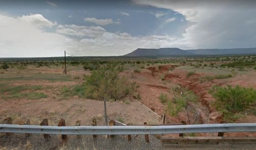 A vast, arid landscape with a distant mountain, cloudy sky, and a dry creek bed, viewed from behind a metal fence.