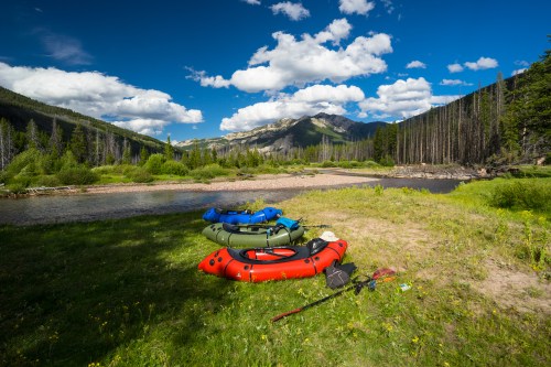 Three colorful inflatable kayaks rest on green grass by a river, surrounded by mountains and blue skies with fluffy clouds.