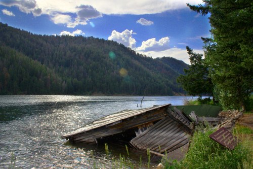 A partially submerged, weathered cabin by a river, surrounded by lush green mountains and a cloudy sky.