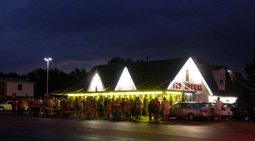 A large crowd gathers outside a brightly lit restaurant at dusk, with cars parked nearby.