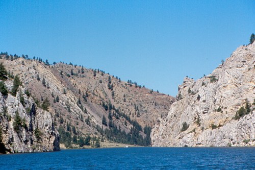 A serene river flows between rocky cliffs and hills under a clear blue sky, surrounded by sparse trees.