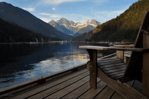 A serene lakeside view with mountains in the background, featuring a wooden dock and a chair overlooking the water.