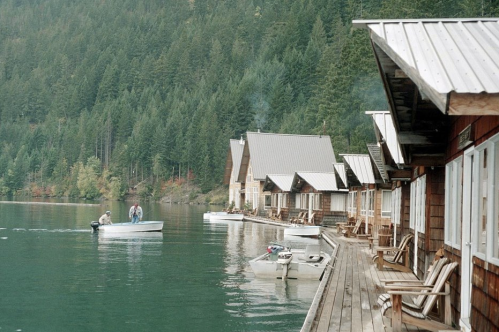 A serene lakeside scene with wooden cabins, a dock, and a small boat with two people on calm water surrounded by trees.