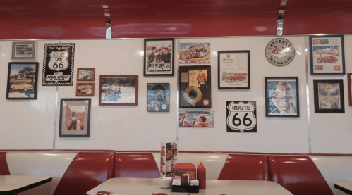 A retro diner wall adorned with vintage photos, Route 66 signs, and memorabilia, featuring red booths and a table.