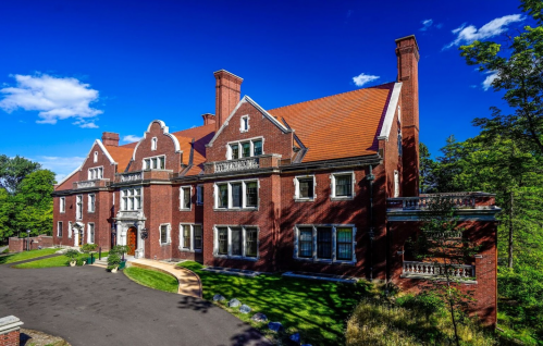 A large, historic brick mansion with a red roof, surrounded by greenery and a clear blue sky.