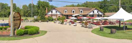A winery with a large building, outdoor seating, and a tent, surrounded by greenery and barrels.