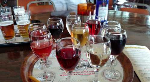 A variety of wine glasses filled with different colored wines on a wooden table, with beer samples in the background.