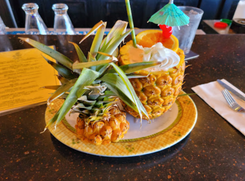 A decorative pineapple drink garnished with an umbrella, orange slice, and whipped cream, alongside a smaller pineapple.