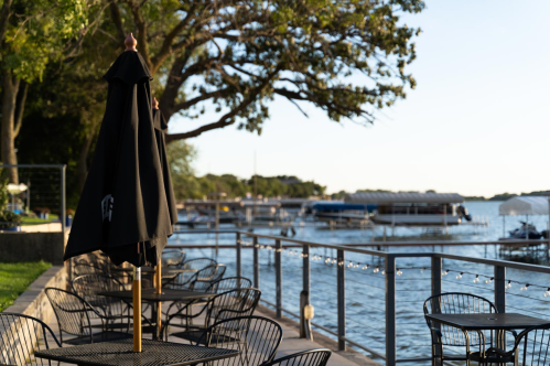 Outdoor dining area by a calm lake, featuring tables with black umbrellas and boats in the background.