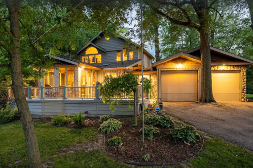 A charming two-story house with a porch, warm lights, and a garage, surrounded by lush greenery.