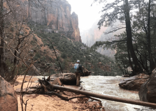 A hiker stands by a river in a canyon, surrounded by rocky cliffs and trees, with mist in the air.