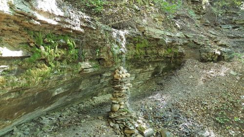 A small stone cairn stands near a rocky cliff, with water trickling down from the top in a serene forest setting.