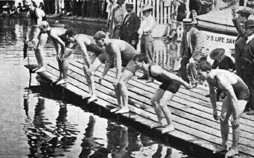 Six male swimmers poised to dive from a wooden dock into a body of water, with spectators watching from the shore.
