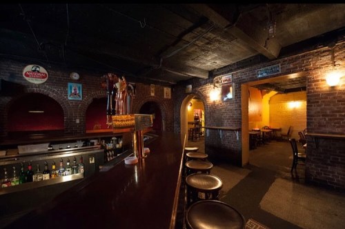 A dimly lit bar with a wooden counter, brick walls, and empty tables in a cozy, inviting atmosphere.