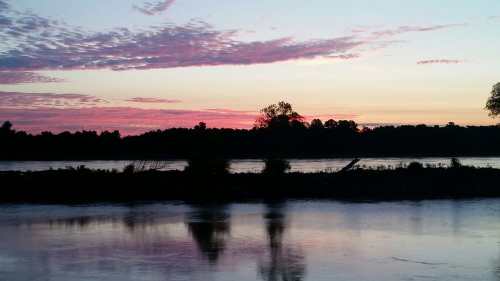 A serene sunset over a river, with pink and purple clouds reflecting on the water's surface.
