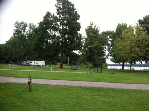 A grassy area with trees, picnic tables, and RVs near a lake under a cloudy sky.