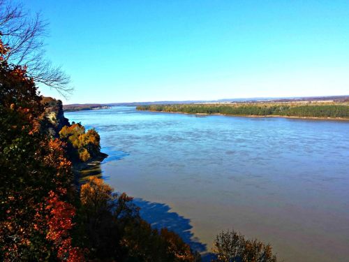 A serene river flows through a landscape of trees, with clear blue skies above and autumn colors along the banks.