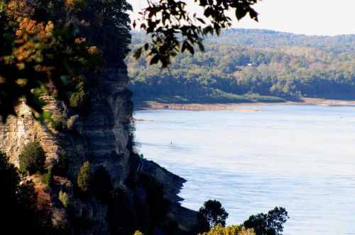 A serene river scene with a rocky cliff on one side and lush greenery along the water's edge.