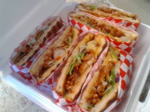 A takeout container filled with several stacked sandwiches, each with layers of meat, vegetables, and red-checkered paper.