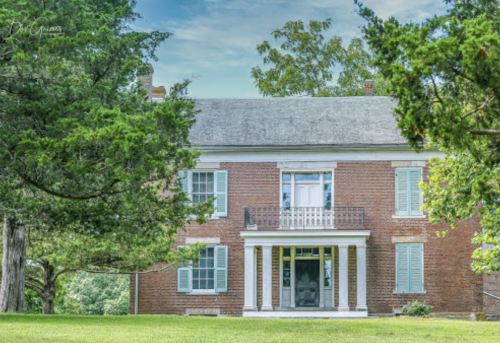 A historic brick house with white shutters and a balcony, surrounded by green trees and a grassy lawn.