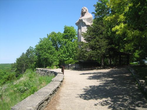 A large statue of a figure stands among trees, overlooking a scenic landscape on a clear day.