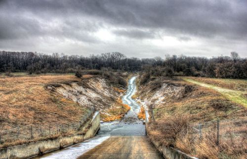 A winding stream flows through a grassy landscape under a cloudy sky, with bare trees in the background.