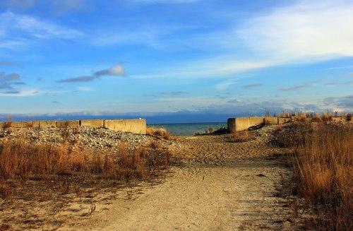A sandy path leads to a calm beach, framed by grass and concrete structures under a blue sky with scattered clouds.