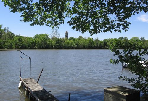 A serene river scene with a wooden dock, lush green trees, and a distant clock tower under a clear blue sky.