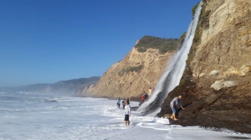 People walking along a beach near a waterfall cascading down a rocky cliff into the ocean under a clear blue sky.