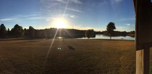A serene landscape featuring a lake at sunset, surrounded by trees and open grass under a clear blue sky.