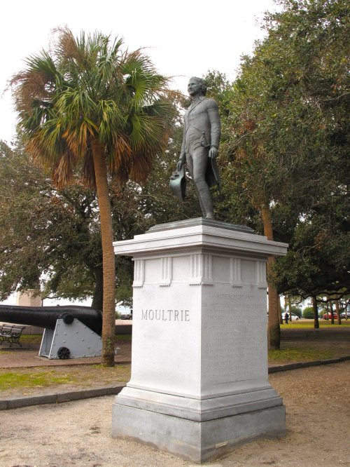 Statue of William Moultrie on a pedestal, surrounded by palm trees and historical cannons in a park setting.