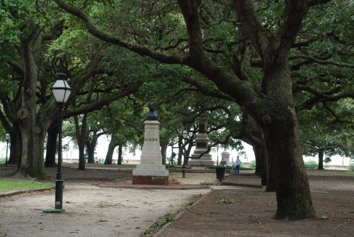 A park scene with trees, lampposts, and statues along a path, creating a serene and shaded atmosphere.