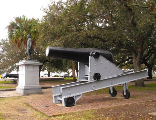 A historic cannon on a wheeled carriage beside a statue in a park with trees and a waterfront in the background.