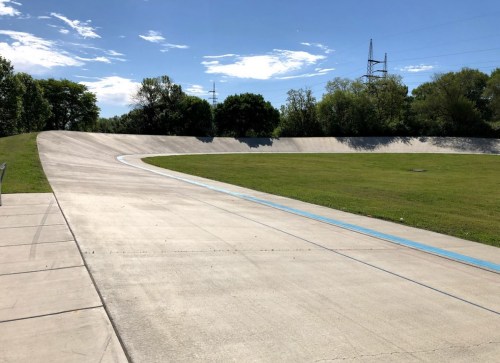 A smooth, curved cycling track surrounded by green grass and trees under a clear blue sky.