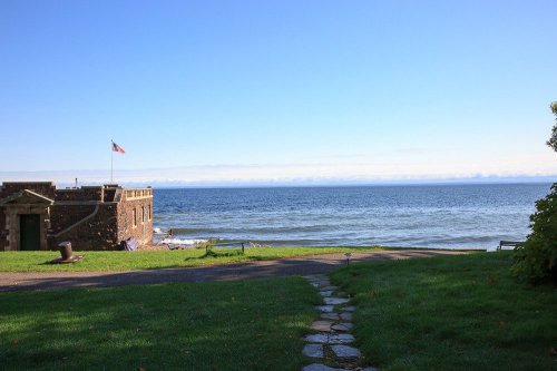 A serene view of the ocean with a stone building and an American flag, framed by green grass and a clear blue sky.